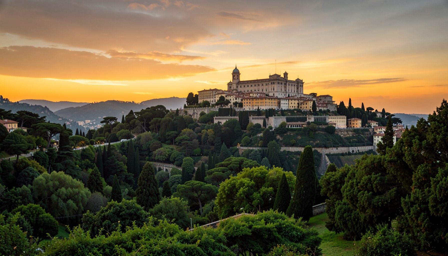 Vista panoramica di un grande complesso collinare tra una fitta vegetazione e cipressi, contro un vivido cielo al tramonto