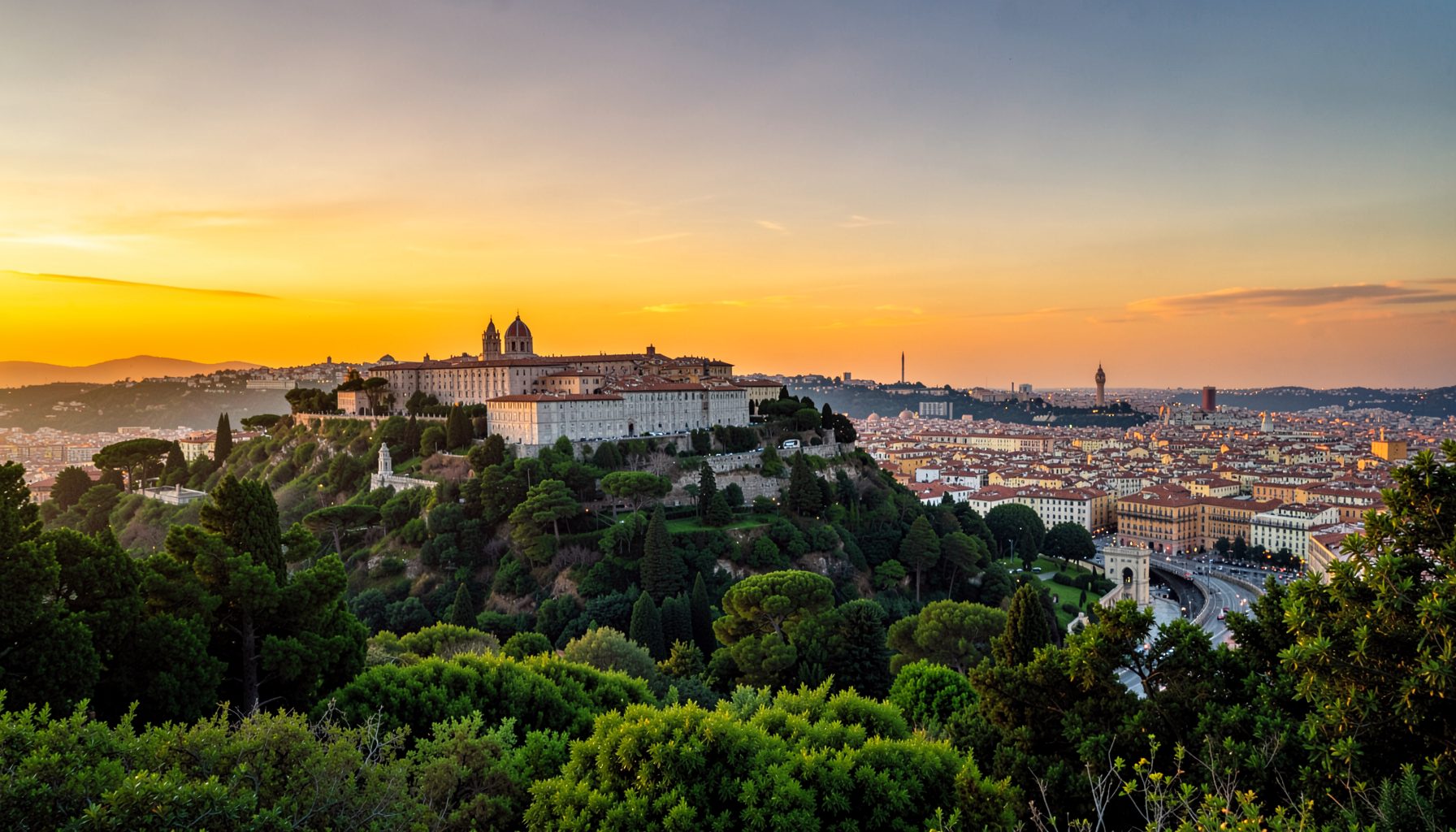 Veduta elevata di un complesso collinare di edifici storici circondato da alberi, che domina una città estesa, sotto un cielo caldo